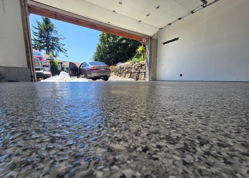 Floor-level view of a finished chip flake garage floor, its high-gloss surface reflecting the open garage door and the driveway beyond